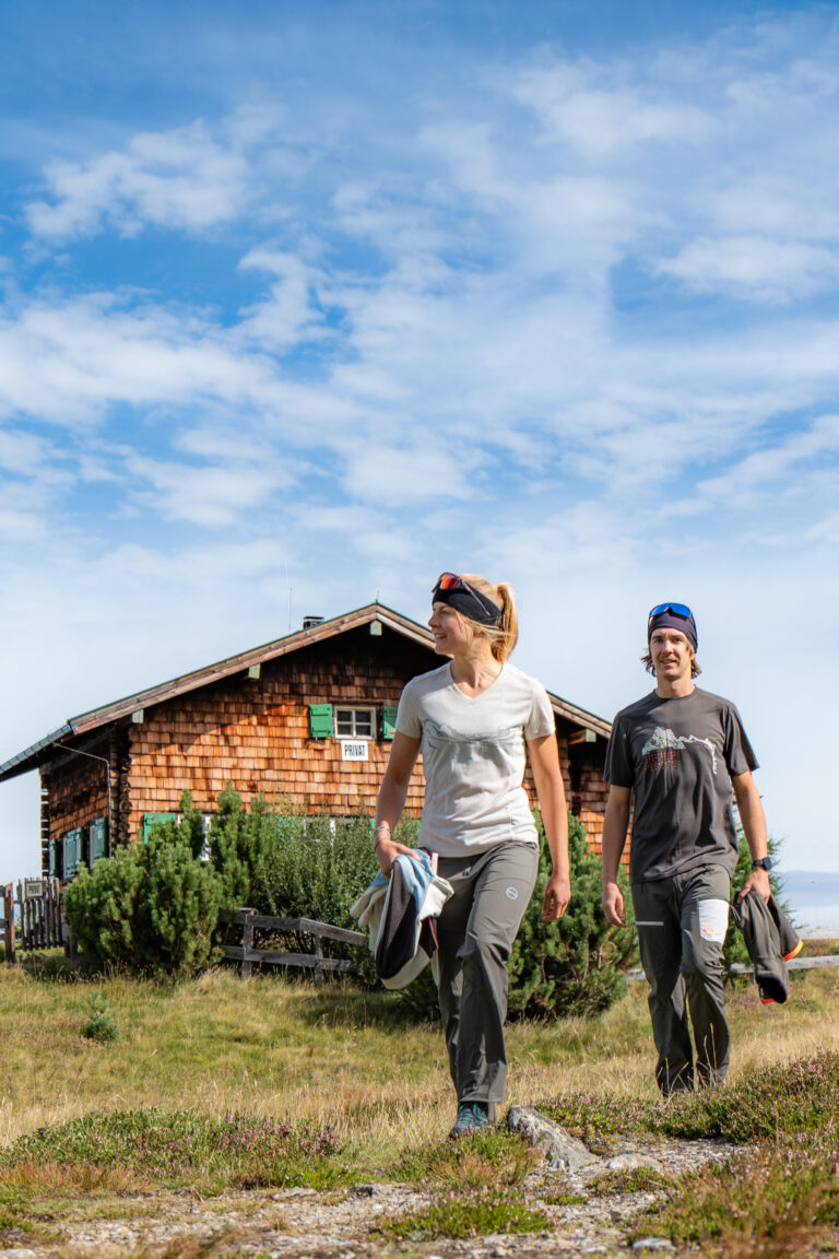 Frau und Mann vor einer Hütte in den Bergen mit strahlend blauem Himmel- mit GTS Sports Kleidung