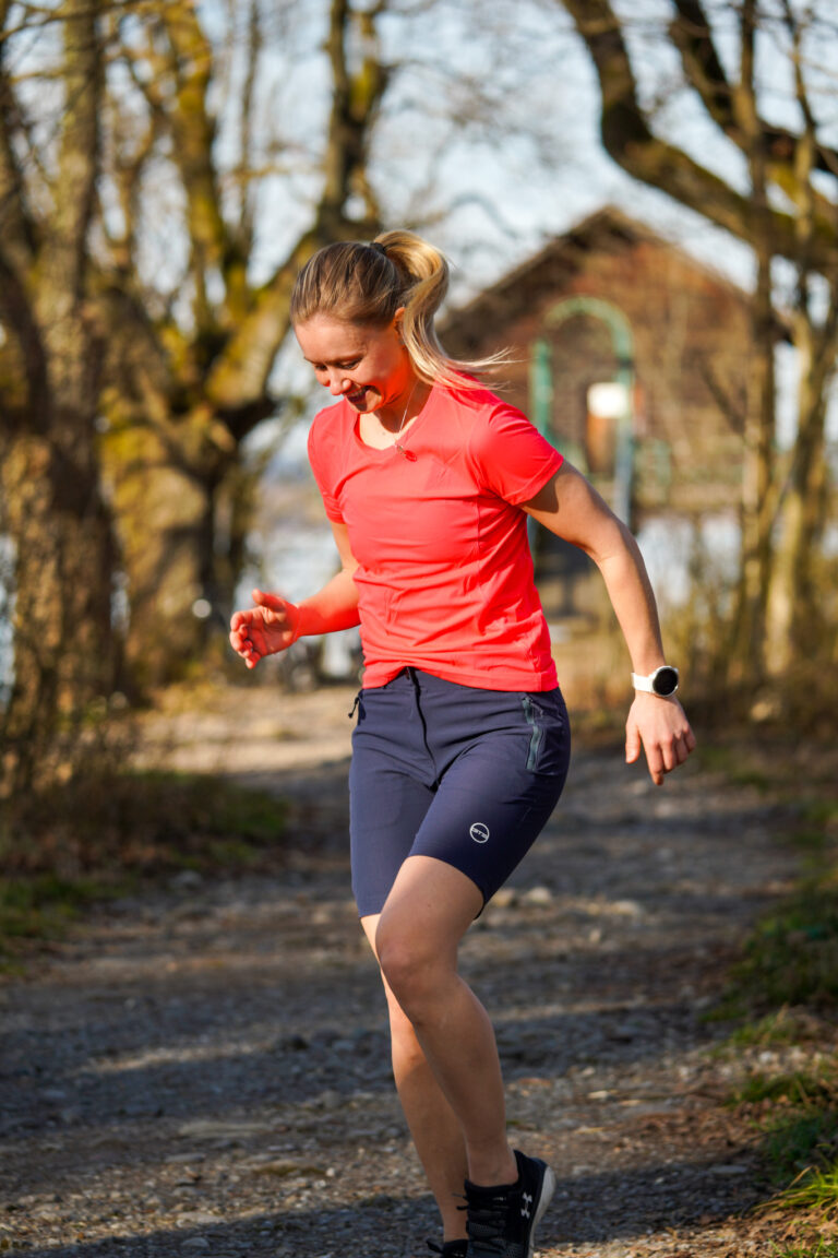 Läuferin macht Übungen auf einem Waldweg und trägt ein neonpinkes GTS Sports Shirt und dunkelblaue Shorts